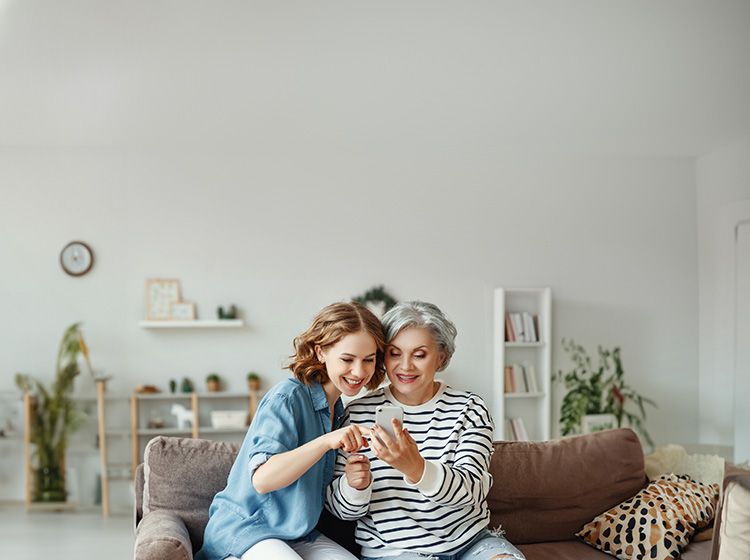 Alte und junge Frau sitzen lachend auf Sofa und schauen auf ein Smartphone