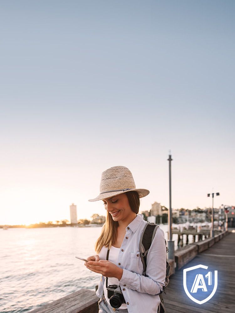 Junge Frau schaut lächelnd auf ihr Smartphone. Im Hintergrund das Meer mit Sonnenuntergang.
