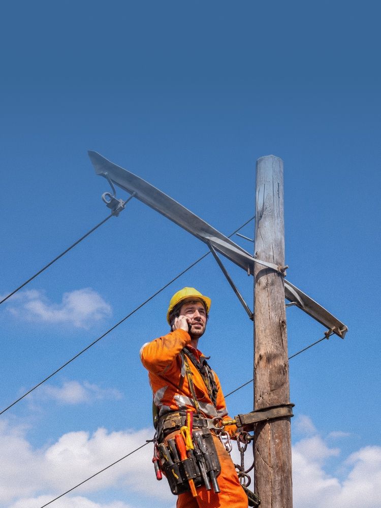 Arbeiter in orangefarbener Schutzkleidung klettert mit Werkzeug an einem Holzmast, blauer Himmel im Hintergrund. Telefoniert über Cloud Telefonanlage mit Handy.