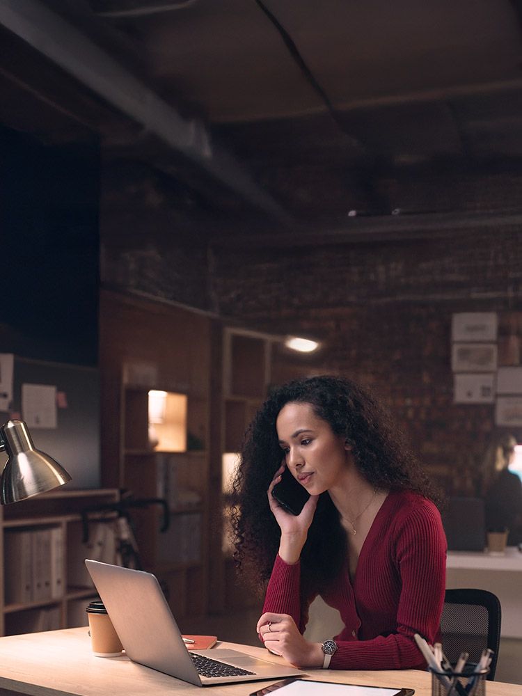 Junge Frau mit dunklen Haaren und roter Jacke sitzt in einem abgedunkeltem Büroraum am Arbeitsplatz vor ihrem offenen Laptop und telefoniert mit einem Handy.