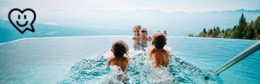 Zwei Kinder schwimmen auf Erwachsene in einem Infinitypool zu, dahinter Berge. Links oben Herzsymbol mit Smiley.