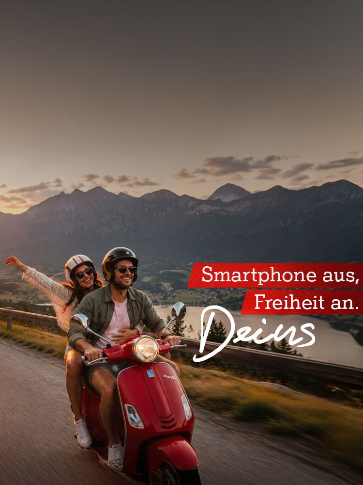 Zwei Personen fahren auf einem roten Roller in den Sonnenuntergang. Text: Smartphone aus, Freiheit an. Deins.