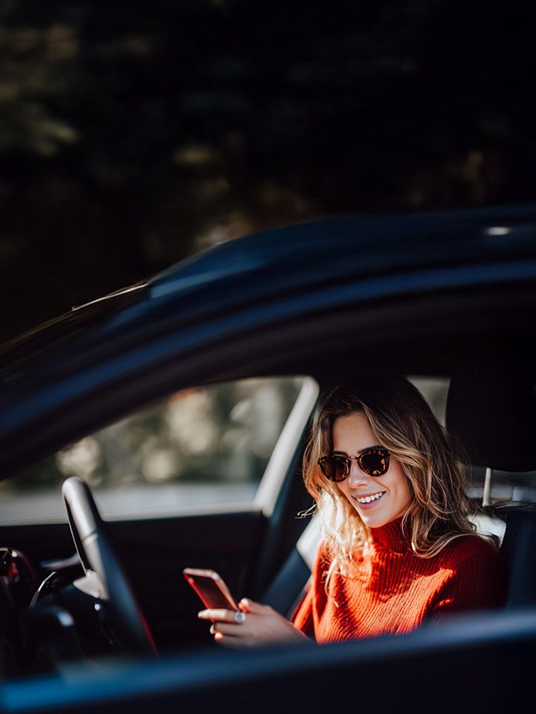 Frau mit rotem Pullover sitzt im Auto und benutzt ein Smartphone.