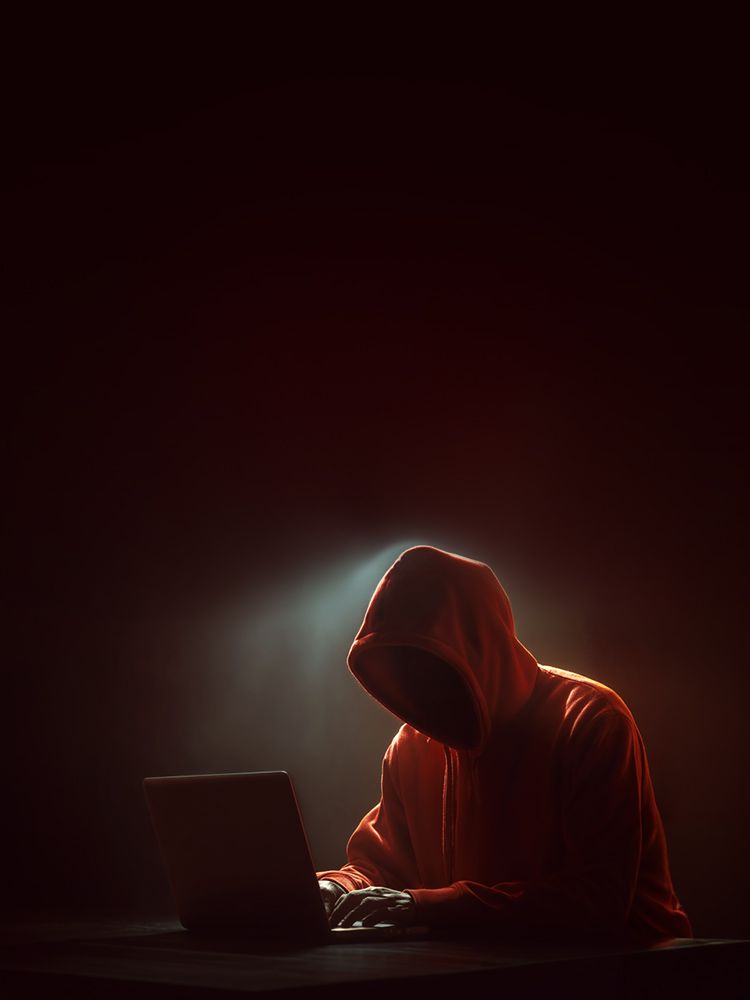 Person mit rotem Kapuzenpullover sitzt im Dunkeln an einem Laptop, Gesicht im Schatten.